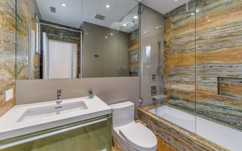 White bathroom sink with green vanity, wall mirror, toilet, and bathtub with yellow stone wall and hand shower